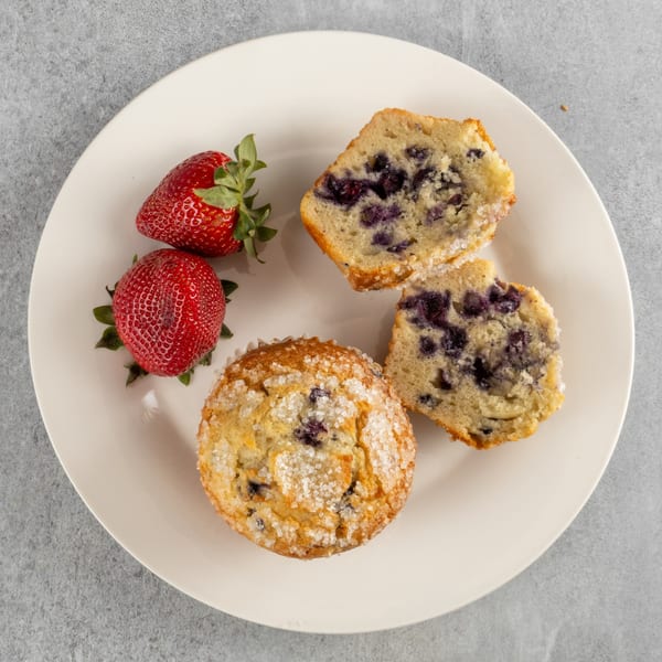 A whole blueberry muffin, two halves of a blueberry muffin, and two fresh strawberries on a white plate.