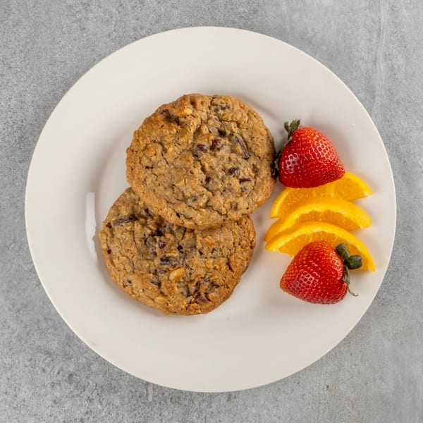 Two oatmeal chocolate chip cookies on a white plate with strawberries and orange slices.
