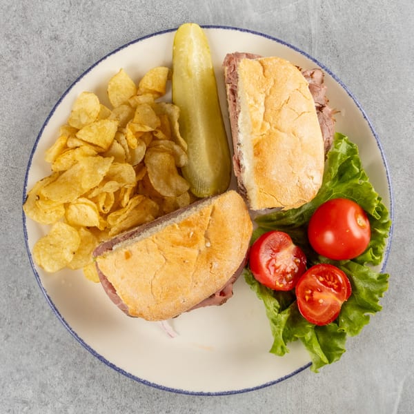 Roast beef sandwich with chips, a pickle spear, and cherry tomatoes on lettuce on a white plate.