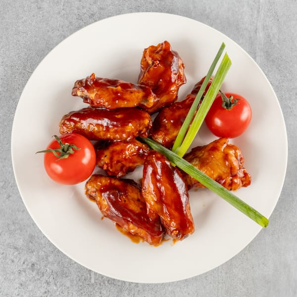 A white plate holds BBQ chicken wings, garnished with two red tomatoes and green onions.