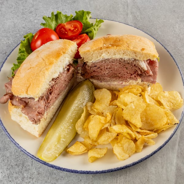 Roast beef sandwich on a white plate with a pickle spear, potato chips, lettuce, and sliced tomatoes.