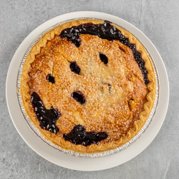 A freshly baked blueberry pie in a foil tin, topped with sparkling sugar, on a white plate.