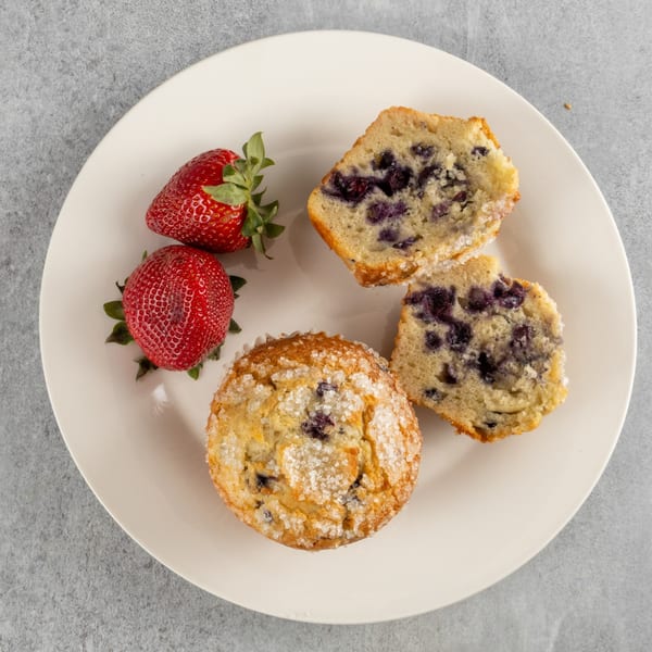 Two fresh strawberries and two blueberry muffins, one whole and one split, on a white plate.
