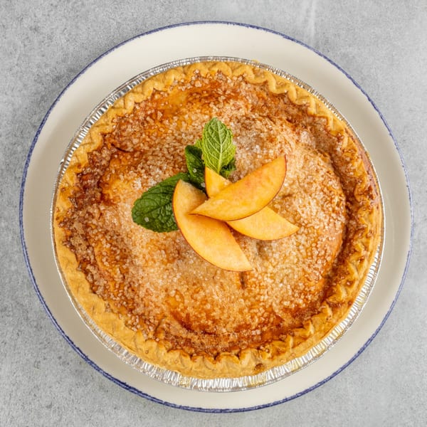 Overhead view of a peach pie in a foil tin, garnished with mint and peach slices.