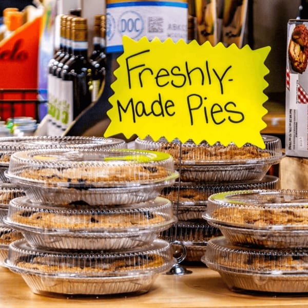 Stacks of freshly made pies in aluminum tins with clear plastic lids, and a yellow sign.